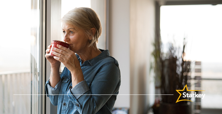 an older woman sips a cup of coffee or tea while looking out the window of her home's living room
